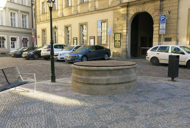 Ein Stadtplatz mit einer Bank in der Mitte, umgeben von Gebäuden, Laternen, Schildern und einem zentralen Springbrunnen.