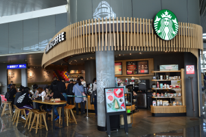 Ein Starbucks-Café in einem Flughafen-Terminal mit Menschen an Tischen und einer Säule mit dem Starbucks-Logo in der Mitte, sowie Brettern an den Wänden und Regalen mit Flaschen auf der rechten Seite.