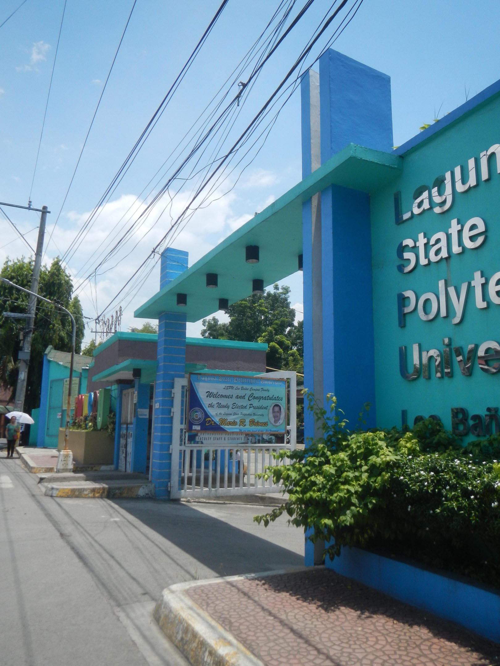 Eingang zum Laguna State Polytechnic University mit einem Namensschild auf der rechten Seite, umgeben von Grün, Gebäuden im Hintergrund und einem klaren blauen Himmel.