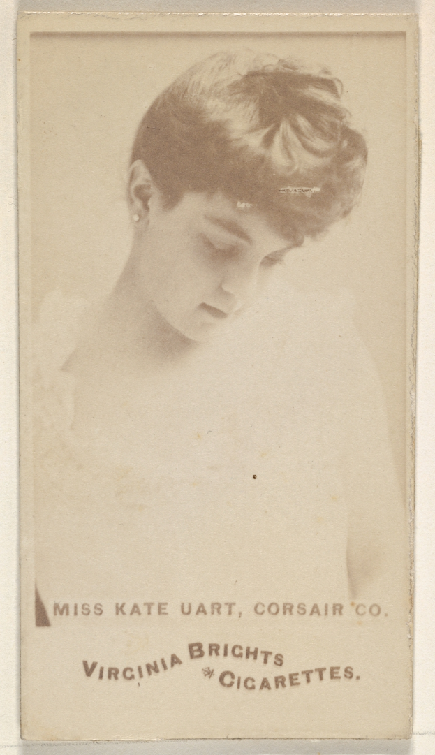 Ein altes Foto einer Frau mit einer Zigarette, die direkt in die Kamera schaut, in einem weißen Kleid und einem gemusterten Schultertuch, mit dem Text "Miss Kate Hart, Corsair Co." unten.