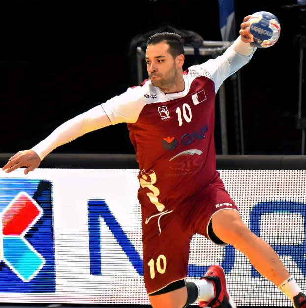 Ein Mann in einer rot-weißen Uniform spielt Handball, hält einen Ball und hat einen entschlossenen Gesichtsausdruck, vor einem Schild mit der Aufschrift "QNP Handball-Weltmeisterschaft 2019".