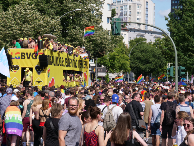 Eine große Menschenmenge marschiert auf einer Straße in Berlin, Deutschland, während eines Christopher Street Day-Umzugs, mit einem gelben Lkw im Hintergrund, viele tragen Mützen und Schutzbrillen, einige halten Fahnen, Laternenmasten, Verkehrsampeln, Bäume und Gebäude mit Fenstern unter einem bewölkten Himmel.