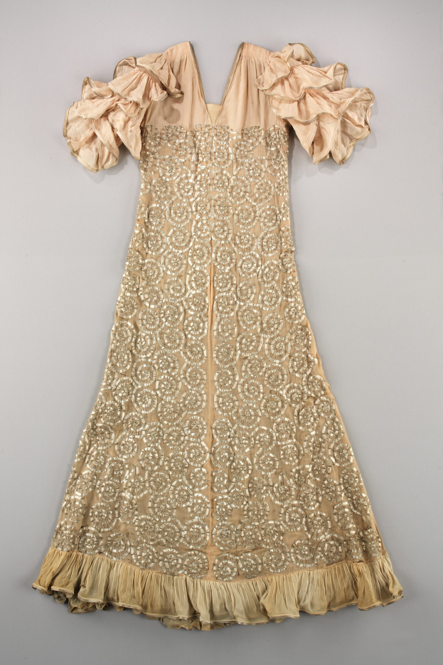 Ein cremefarbenes Kleid mit goldener Stickerei und Rüschen an den Ärmeln, das auf einer weißen Oberfläche präsentiert wird und mit detaillierten Designs und Mustern verziert ist.