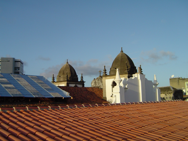 Stadtlandschaft mit mehreren Gebäuden im Vordergrund und einem klaren blauen Himmel im Hintergrund, mit Solarpanelen auf einem Dach, die den Einsatz erneuerbarer Energien anzeigen.