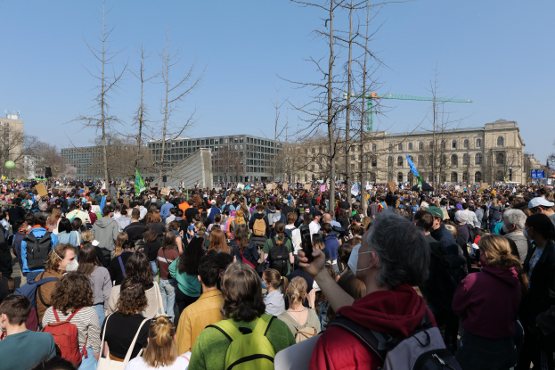 Eine große Menschenmenge mit Schildern und Taschen steht vor einem Gebäude mit Fenstern, Bäumen und einem klaren Himmel während einer Klimaprotest.