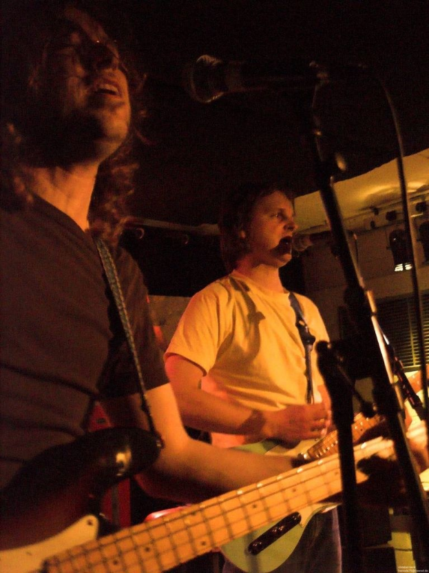 Ein Musikkonzert mit zwei Sängern: einer in einem weißen Hemd mit einer grünen Gitarre und einer in einem schwarzen Hemd mit Brille und einer schwarzen Gitarre, beide performen mit Mikrofonen und anderen Instrumenten im Hintergrund.