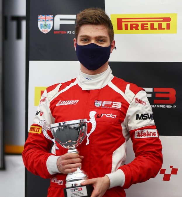 Ein Mann in einem roten und weißen Rennanzug und einer Gesichtsmaske hält einen Pokal, mit einer schwarzen und weißen Tafel und einem gerahmten Gegenstand im Hintergrund.