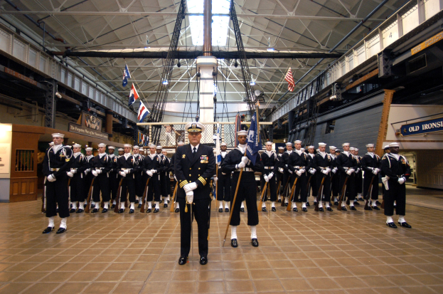 Eine Gruppe uniformierter Männer in einem großen Raum, die Mützen tragen und Gewehre halten, mit Fahnen und Schildern im Hintergrund.