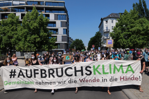 Gruppe von Menschen mit Masken, die ein Banner mit der Aufschrift "Aufbruchsklima" tragen, vor einem Gebäude mit Bäumen und klarem Himmel.