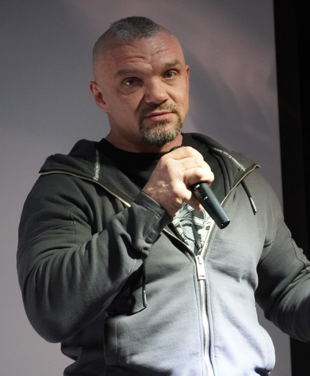 Ein Mann in einer schwarzen Kapuzenjacke und einem schwarzen Anzug mit weißem Hemd hält ein Mikrofon und steht selbstbewusst vor einer Wand, wobei er wie ein Komiker wirkt.