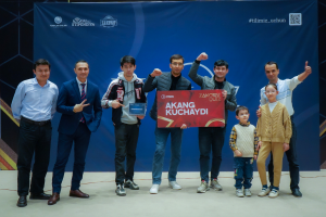 Eine Gruppe von Menschen auf einer Bühne, zwei halten ein Schild mit dem Text 'Akang Kuchaydi - Zakovat Quiz', mit einem Banner, Lichtern und anderen Gegenständen im Hintergrund.