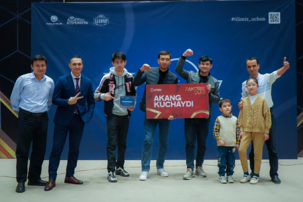 Eine Gruppe von Menschen auf einer Bühne, zwei halten ein Schild mit dem Text 'Akang Kuchaydi - Zakovat Quiz', mit einem Banner, Lichtern und anderen Gegenständen im Hintergrund.