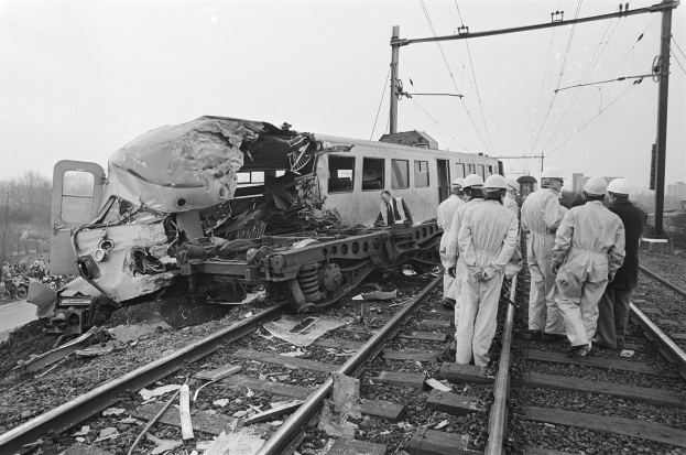Eine Schwarz-Weiß-Szene eines Zugunfalls mit Menschen in Helmen drumherum, Strommasten, Bäume, Gebäude und einen klaren Himmel im Hintergrund.