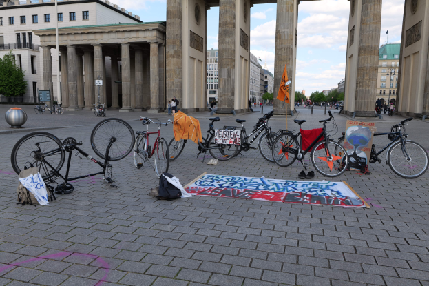 Eine Gruppe von Fahrrädern, die vor dem Brandenburger Tor geparkt sind, mit Taschen und einer Fahne in der Nähe, vor einer Kulisse aus Gebäuden, Bäumen und einem klaren blauen Himmel.
