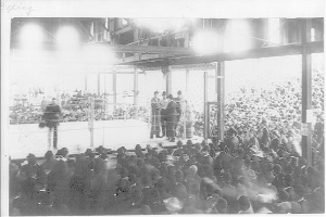 Ein Schwarz-Weiß-Foto von einer Menschenmenge bei einem Boxkampf in einer Arena, mit einigen Individuen auf der Bühne und anderen im Publikum sitzend.