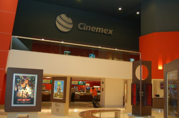 Eine Gruppe von Menschen steht in einem Cinemex-Kino in einem Einkaufszentrum, mit einer Person auf einem Stuhl, Tischen, Fotorahmen an der Wand, einem Schild, Deckenlampen und Geräten auf den Tischen.