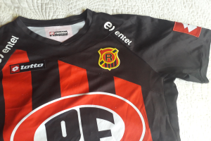 Ein schwarzes und rotes Fussballtrikot mit einem roten und schwarzen Streifen, einem Logo und Text auf einer weissen Fläche.