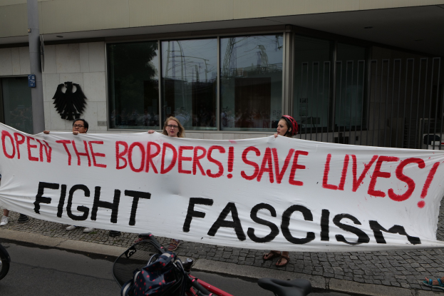Gruppe von Menschen mit einem Transparent mit der Aufschrift "Öffnet die Grenzen, rettet Leben, kämpft gegen Faschismus" vor einem Gebäude mit Glasfenstern und einem Mast, mit einem Fahrrad und einer Tasche auf der Straße.