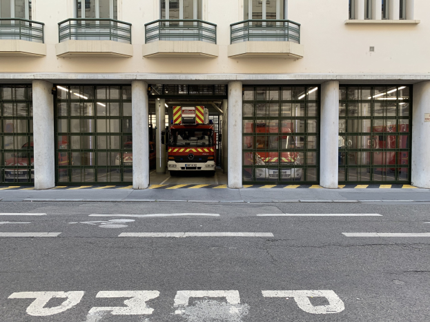 Feuerwache mit Feuerwehrauto davor, mit Fenstern, Geländern, Säulen und Glastüren, sowie einer Straße unten.