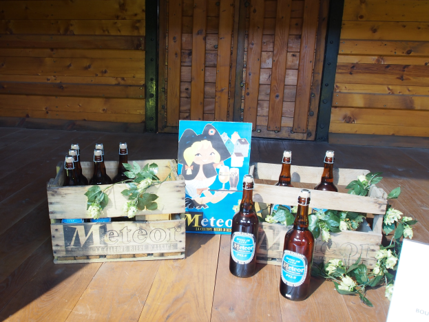 Eine hölzerne Kiste voller Bierflaschen auf einem Holzboden in einer Gartenumgebung, mit Blättern und Blumen in der Nähe, vor einer hölzernen Wand mit einer Tür im Hintergrund.