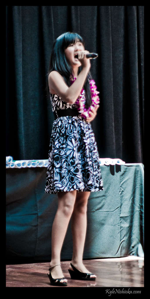 Eine Frau in einem farbenfrohen Kleid singt in ein Mikrofon auf der B├╝hne, mit einem Tisch mit Papieren und einem Vorhang im Hintergrund.