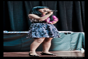 Eine Frau in einem farbenfrohen Kleid singt in ein Mikrofon auf der B├╝hne, mit einem Tisch mit Papieren und einem Vorhang im Hintergrund.