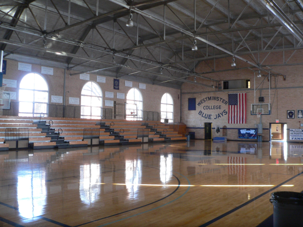 Großer Gymnastikraum mit Holzboden, Basketballfeld, Treppen mit Geländern, Wand mit Fenstern, Texttafeln, Flagge, Deckenleuchten, Basketballkorb und verstreuten Gegenständen.