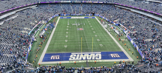 Ein volles Fußballstadion mit Spielern auf dem Feld, Torpfosten und Texttafeln, umgeben von leeren Sitzplätzen.