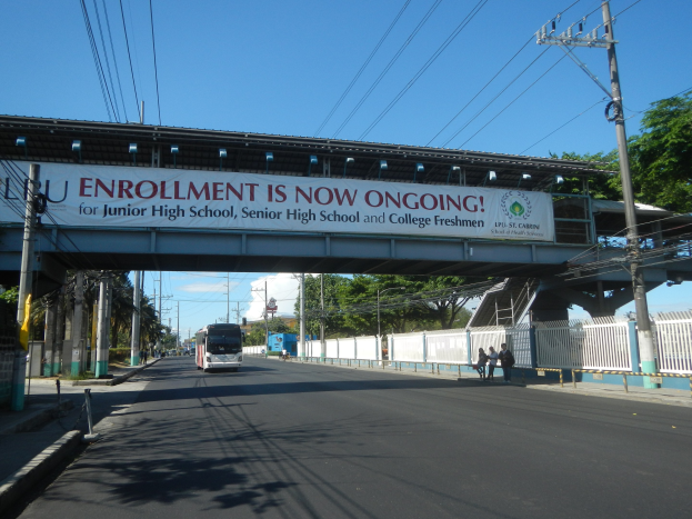 Ein Bus fährt unter einer Brücke mit einem Schild, auf dem "Jetzt ist Einschreibung" steht, während Fußgänger auf dem Gehweg darunter gehen. Elektromasten mit Drähten säumen die Straße, und im Hintergrund sind Bäume und ein klarer blauer Himmel zu sehen.