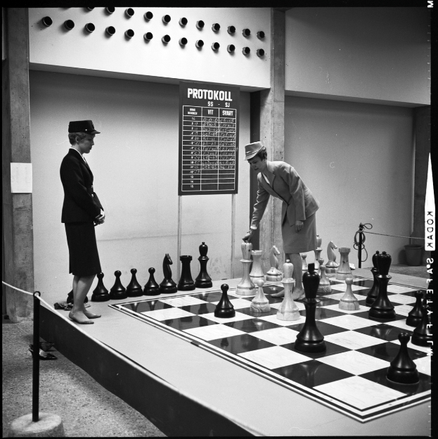 Zwei Frauen spielen Schach auf einem großen schwarz-weißen Brett vor einer Wand mit einer Tafel daran, mit Schachfiguren auf dem Brett.