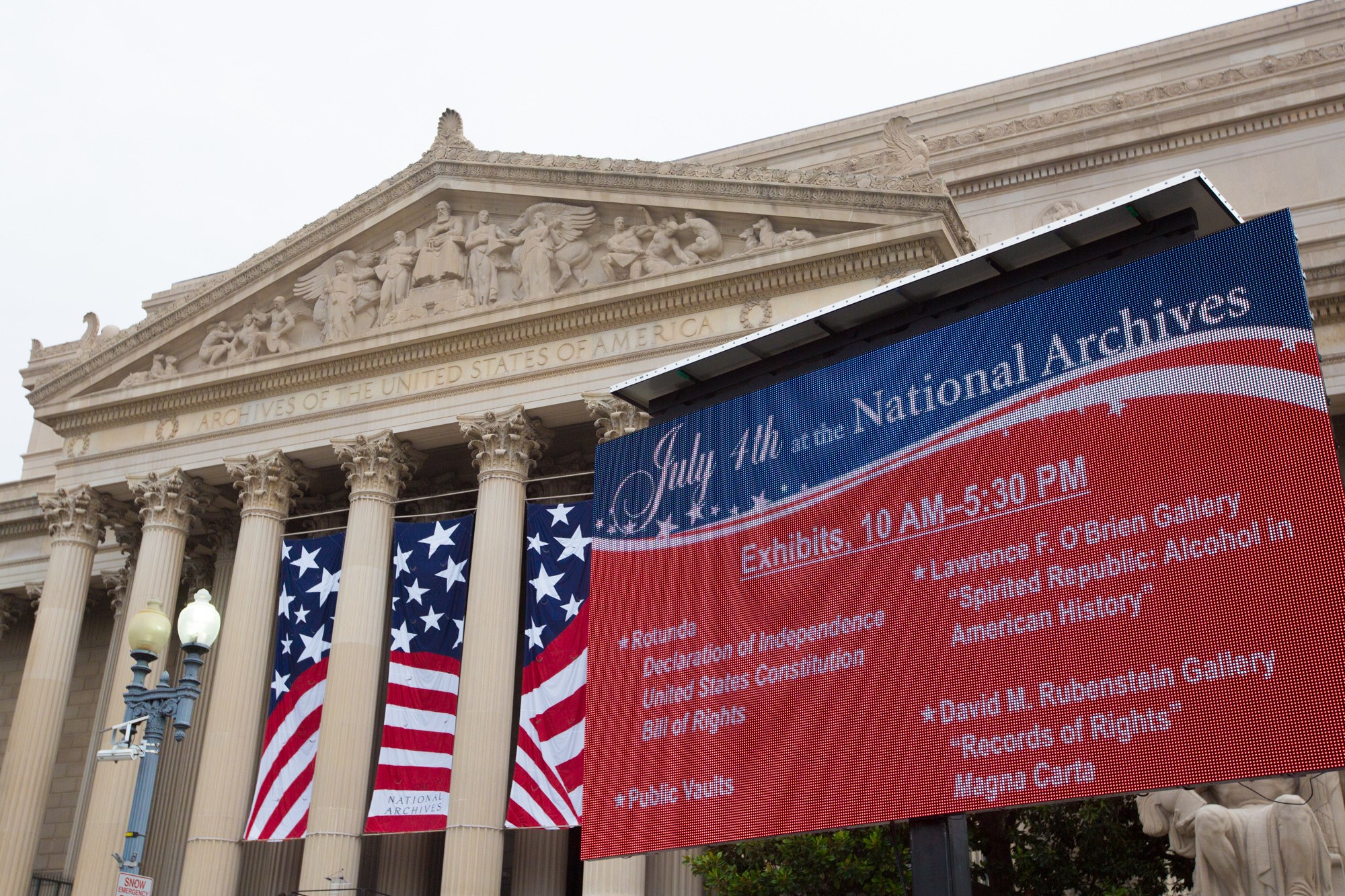 Außenansicht des National Archives-Gebäudes in Washington, DC, mit einer Fahne, Flaggen, Laternenmasten, Bäumen und Skulpturen unter einem klaren blauen Himmel.