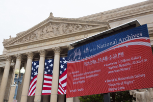 Außenansicht des National Archives-Gebäudes in Washington, DC, mit einer Fahne, Flaggen, Laternenmasten, Bäumen und Skulpturen unter einem klaren blauen Himmel.