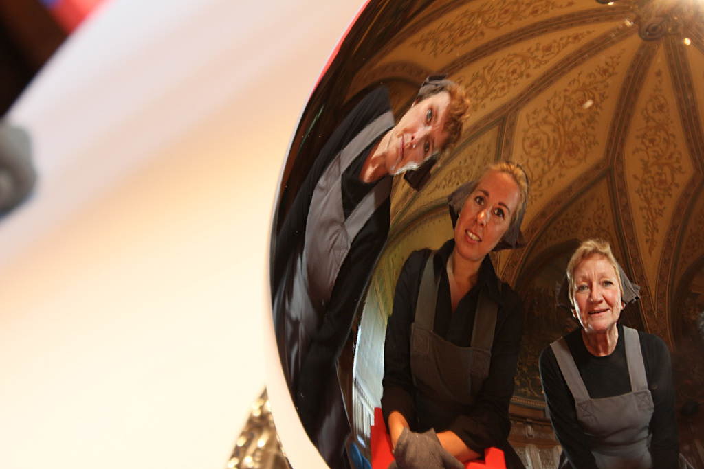 Ein reflektierender Ball auf einem weißen Tisch zeigt das Spiegelbild von drei lächelnden Frauen, die leicht gebeugt sind, mit einem Dach im Hintergrund.