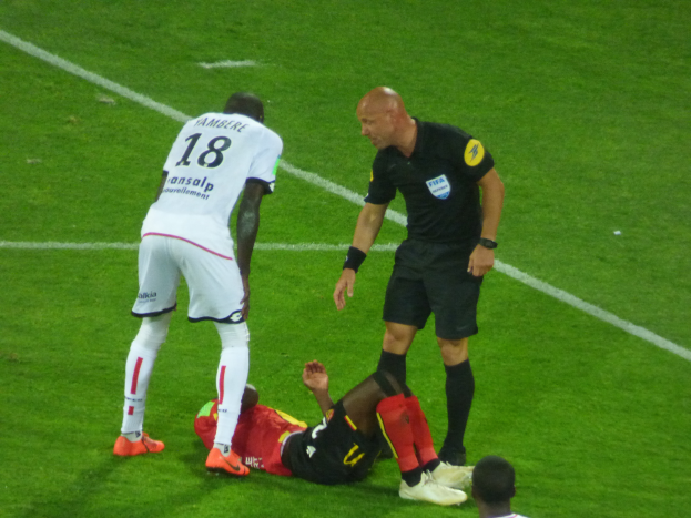Ein Fußballspieler liegt neben einem Schiedsrichter auf dem Boden, umgeben von drei stehenden Personen auf einem Rasenfeld, wobei eine zentrale Figur eine entschlossene Miene und ausgestreckte Arme zeigt.