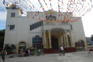 Eine Gruppe von Menschen auf Fahrrädern vor einer weißen Kirche mit architektonischen Details wie Fenstern, Säulen, Bögen, Türen und einem Kreuzsymbol, mit Pflanzen, einem Zaun, einer Fahne, Bäumen und einem bewölkten Himmel im Hintergrund und bunten Fahnen, die an einem Seil über ihnen befestigt sind.