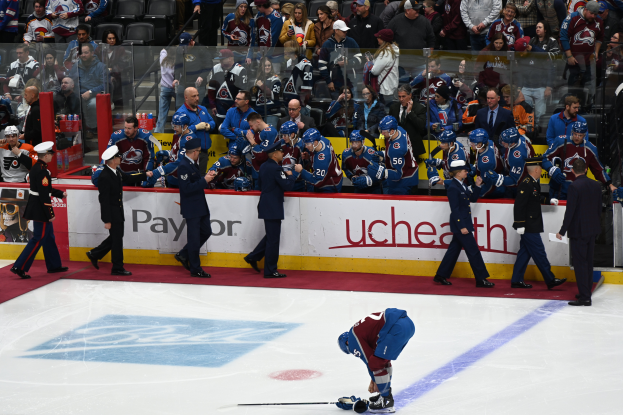 Ein Hockey-Spieler auf dem Eis mit einem Stock in der Hand, umgeben von einer Gruppe von Menschen mit Mützen und Hockey-Stöcken, mit Zuschauern und Brettern im Hintergrund, auf denen "Colorado Avalanche vs San Jose Sharks - 3/24/15 NHL Pick, Odds, and Prediction" steht.
