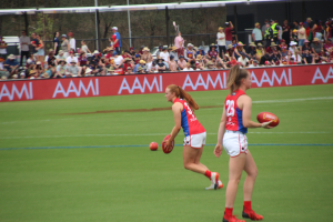 Zwei Frauen in Sportbekleidung spielen AFL-Fußball auf einem Feld, eine hält den Ball, mit Zuschauern und Veranstaltungsbeschilderung im Hintergrund.