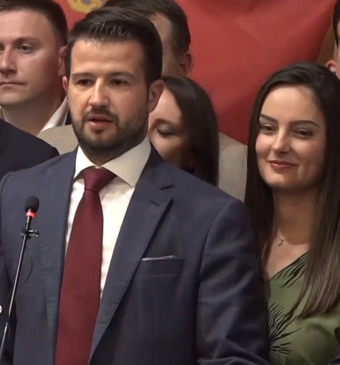 Mann im Anzug und Krawatte spricht vor Publikum mit Mikrofon, mit einem Banner im Hintergrund.