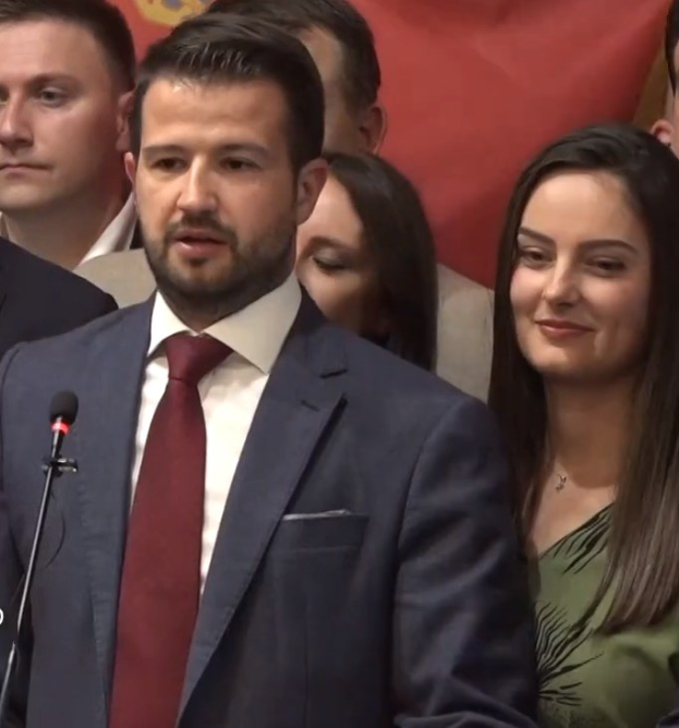 Mann im Anzug und Krawatte spricht vor Publikum mit Mikrofon, mit einem Banner im Hintergrund.