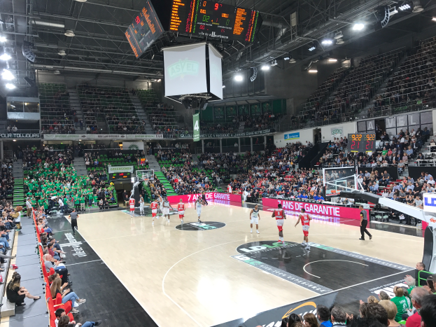 Ein Basketballspiel in einer gro├čen Arena mit Zuschauern auf den R├Ąngen, Anzeigetafeln mit "Asvel vs Galatasaray" und einer Decke mit Lichtern und einem Bildschirm.
