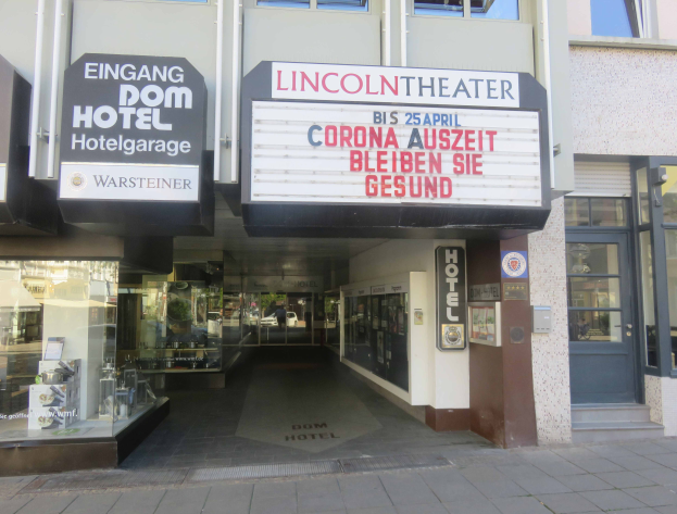 Das Lincoln Theater in Berlin, Deutschland, ein Gebäude mit Glasfenstern und -türen und einer Tafel mit Text darauf, das verschiedene Gegenstände im Inneren zeigt und den Eindruck einer pulsierenden Stadtlandschaft vermittelt.