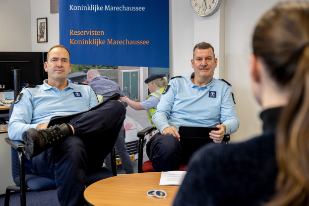 Zwei Polizeibeamte sitzen an einem Tisch mit einer Frau, umgeben von Dokumenten und Büromaterialien, mit einer Wandbanner, Uhr und Fotorahmen im Hintergrund und einem zweiten Tisch mit einem Computer-Setup links.