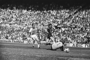 Schwarzes Foto von Männer, die auf einem Feld Füßball spielen, mit Zuschauern im Hintergrund und einem Banner 'Bundesliga 1978-79 Ajax 1-1 Hannover 96'.