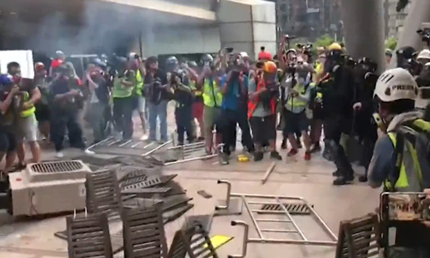 Menschen in Helmen und Jacken stehen vor einem raucherfüllten Gebäude, einige halten Kameras, mit verstreuten Metallobjekten und Stadtpfeilern im Hintergrund.