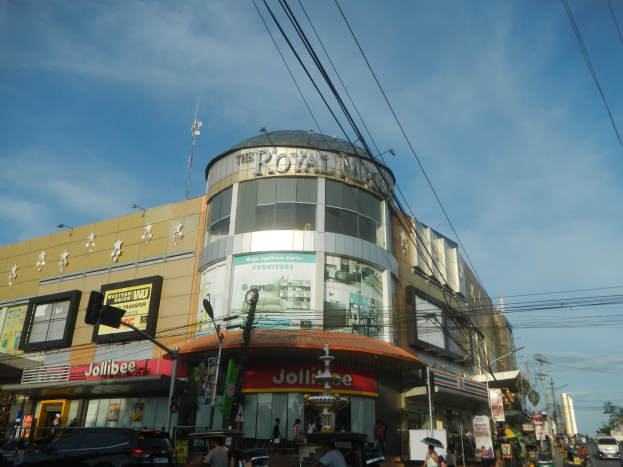 Eine belebte Straße mit fahrenden Fahrzeugen, Fussgängern auf dem Gehweg und einem großen Gebäude mit der Aufschrift "Royal Mall" im Hintergrund, umgeben von parkenden Autos, Bäumen, Strommästen und Drähten unter dem Himmel.