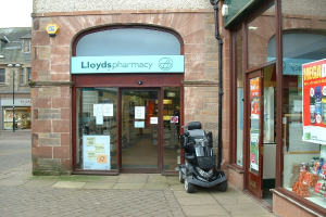 Eine Apothekenfiliale mit einem davor geparkten Fahrzeug und einem im linken Eck sichtbaren Gebäude.