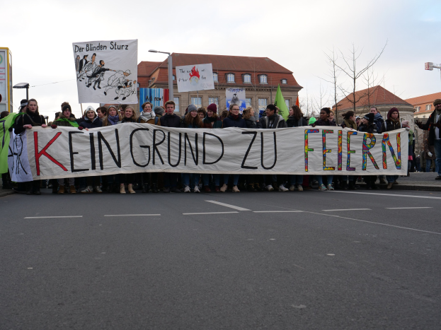 Protestierende mit einem Banner "Kein Grund zu Feiern" gegen deutsche Sparmaßnahmen, mit Gebäuden, Bäumen und einem klaren Himmel im Hintergrund.