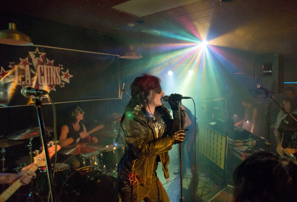 Eine Frau in einer schwarzen Jacke singt in ein Mikrofon auf der Bühne, mit einem Mann in einer Sonnenbrille und einem Stirnband, der ein Instrument spielt, und einem Plakat mit 'LA Guns' im Hintergrund.