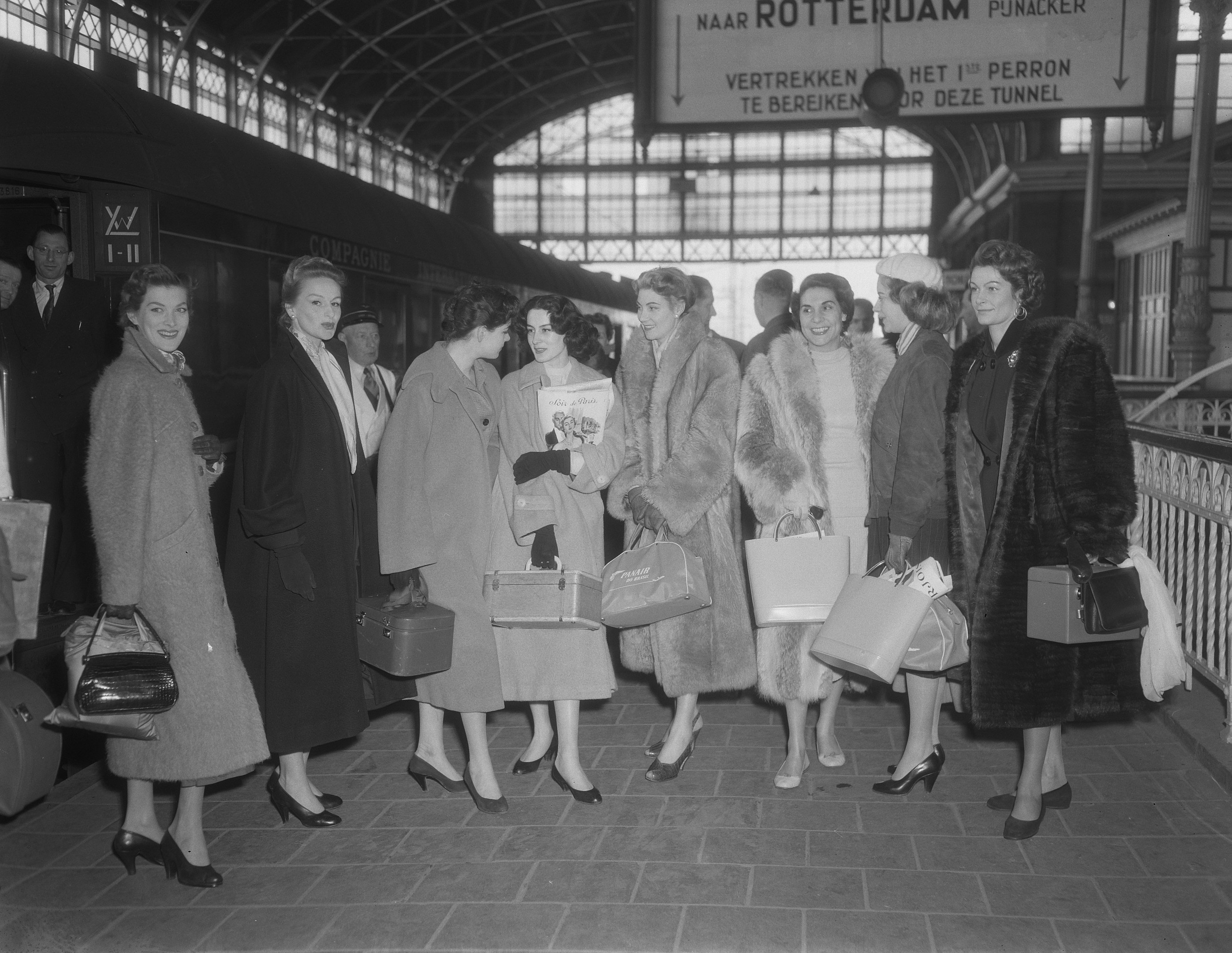 Schwarz-weißes Bild von Frauen, die an einem Bahnhof mit Taschen stehen, neben einem Zug auf der linken Seite und Geländern mit einer Tafel auf der rechten Seite.
