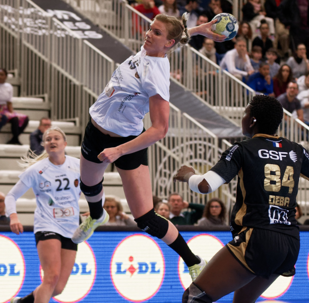 Eine Gruppe von Frauen, die auf einem Platz Handball spielen, mit einem Ball in der Luft, während Zuschauer von hinten zusehen, wo ein "Handball-Weltmeisterschaft 2019 - Tag 2"-Schild sichtbar ist.